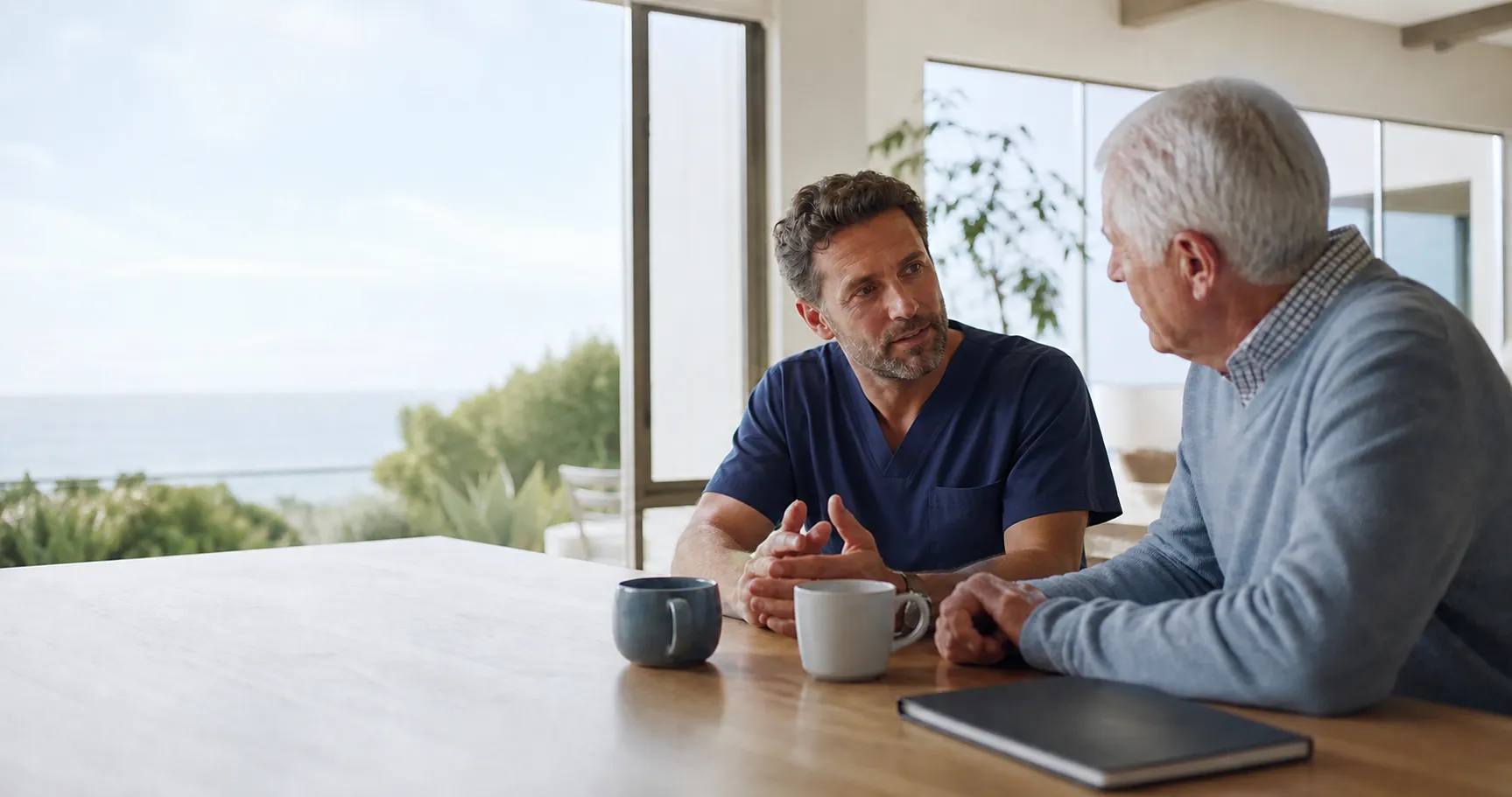 A physician in navy scrubs speaks with an older man at a table
